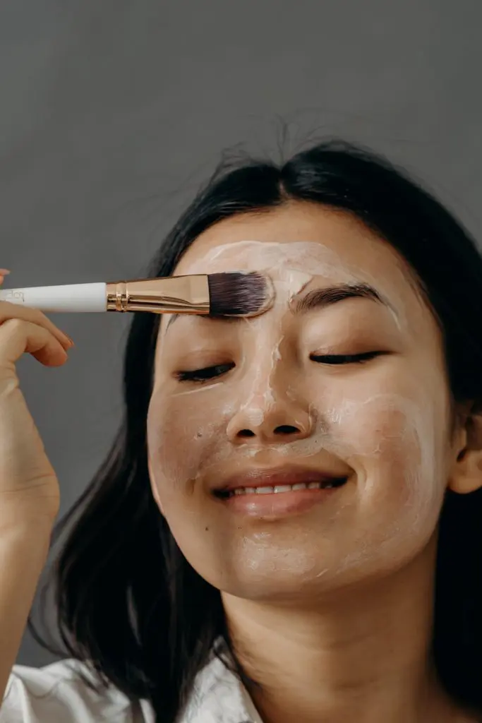 Donna asiatica con pelle luminosa che applica un prodotto skincare davanti a uno specchio, circondata da cosmetici coreani su sfondo minimalista e chiaro. Rappresentazione della routine di bellezza coreana passo dopo passo